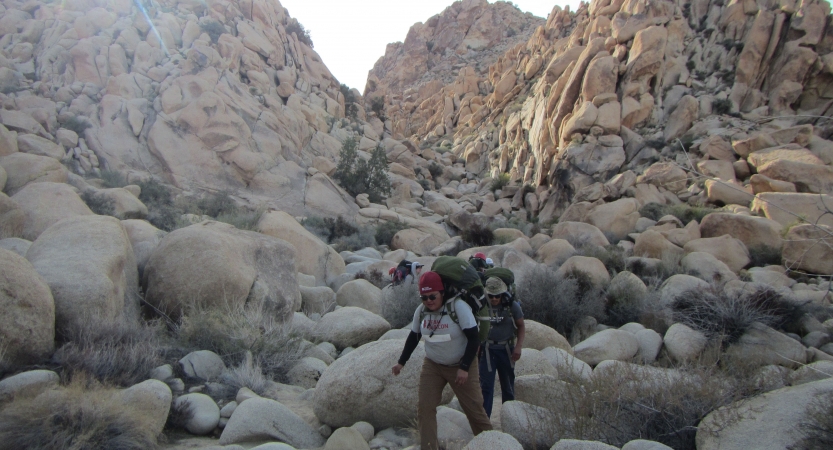 A group of people wearing backpacks make their way through an area populated by large rocks.